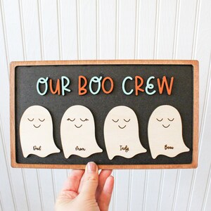May include: A wooden sign with the text "Our Boo Crew" in orange and blue letters. Four wooden ghost shapes are attached to the sign, each with a name written on it: Dad, Mom, Indy, and Beau.