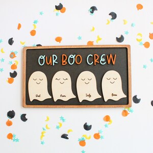 May include: A wooden sign with a black chalkboard background and a wooden frame. The sign says "Our Boo Crew" in orange and blue letters. There are four white ghost cutouts with the names "Dad", "Mom", "Indy", and "Beau" written on them.