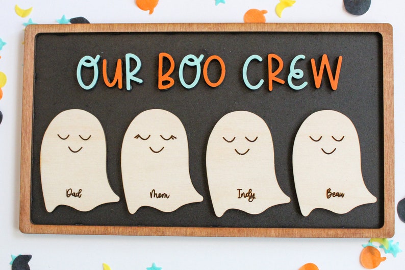 May include: A wooden sign with the text "Our Boo Crew" in orange and blue letters. There are four ghost-shaped cutouts with the names "Dad", "Mom", "Indy", and "Beau" written on them.