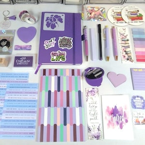 May include: A collection of stationery items including a purple notebook with stickers, a multi-colored striped notepad, various pens, bookmarks, a craft ink pad, heart-shaped cutouts, adhesive tabs, and circular stickers with the text There is no such thing as too many books.