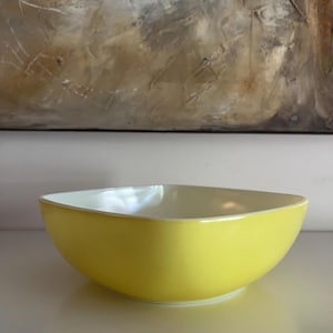 Puede incluir: Un cuenco cuadrado de cerámica amarilla con interior blanco. El cuenco tiene un acabado liso y brillante y una base ligeramente redondeada. El cuenco está vacío y se encuentra sobre una superficie blanca.