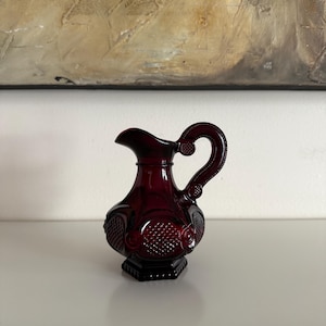 Könnte beinhalten: Kleine, rubinrote Glaskanne mit dekorativem Griff und Ausguss. Die Kanne hat ein verziertes, strukturiertes Design am Körper und steht auf einem sechseckigen Sockel. Wahrscheinlich Vintage oder Antik.