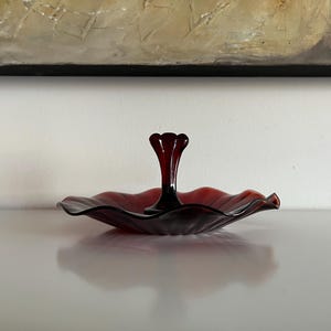May include: A dark red glass serving dish with a central stem. The dish has a scalloped edge and a glossy finish. The stem is a deep red colour. The dish is likely used for serving small portions of food or displaying decorative items.