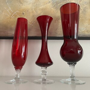 May include: Three red glass vases with clear glass bases. The vases are different shapes and sizes. The tallest vase has a round bulb at the bottom and a flared top. The middle vase has a narrow neck and a wide base. The shortest vase has a straight neck and a flared base.