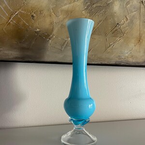 Puede incluir: Un jarrón alto de cristal turquesa con una sección media bulbosa y una parte superior acampanada. El jarrón se asienta sobre una base de pedestal transparente. El fondo presenta una pintura abstracta texturizada con tonos marrones y dorados.