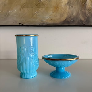Puede incluir: Un jarrón de cristal azul claro y un cuenco pequeño con pedestal y ribete dorado. El jarrón tiene un diseño ornamentado y el cuenco tiene un borde festoneado. Ambos artículos están sobre una superficie blanca.