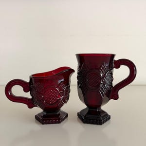 May include: A deep red glass creamer and a matching tall mug, both featuring a textured geometric pattern with circular and diamond motifs. Each piece stands on a hexagonal base and includes a curved handle with a detailed, ornate design.