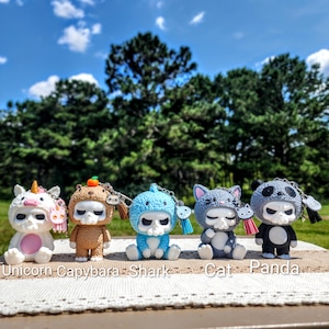 May include: A collection of five whimsical skull figurines, each adorned with a different animal-themed costume: a unicorn, capybara, shark, cat, and panda. The figurines are white with black accents and are sitting on a white lace tablecloth.