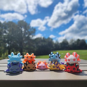 May include: A collection of eight colorful axolotl figurines, each with a unique design and pose. The figurines are made of a smooth, plastic-like material and are arranged in a row on a table.