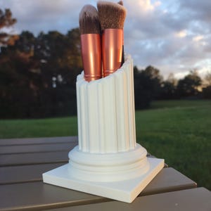 Könnte beinhalten: Weißer Keramik-Make-up-Pinselhalter in Säulenform mit quadratischem Sockel. Der Halter hat ein geriffeltes Design und hält drei Make-up-Pinsel mit roségoldenen Griffen.