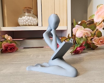 May include: A gray, abstract sculpture of a person sitting and reading a book. The sculpture is made of a smooth, matte material and has a simple, modern design.