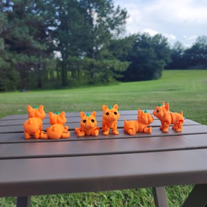 Könnte beinhalten: Sechs orangefarbene, kürbisförmige Hundefiguren in verschiedenen Posen. Die Figuren sind in einer Reihe auf einem grauen Tisch angeordnet.