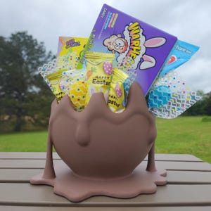 Puede incluir: Un contenedor de chocolate marrón en forma de huevo de Pascua con un efecto de chocolate derretido. El contenedor está lleno de varios dulces de Pascua, incluyendo caramelos agrios Warheads, Sour Patch Kids y otros dulces surtidos. Los dulces están envueltos en celofán de colores con diseños de Pascua.