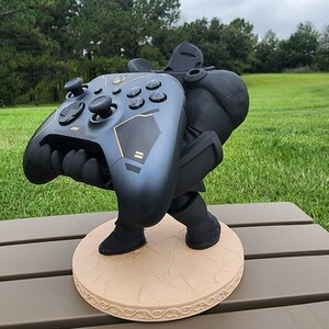 Può includere: Un supporto per controller di videogiochi nero e oro a forma di robot che tiene il controller. Il supporto è su una base beige con una superficie testurizzata.