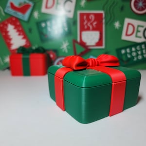 Pode incluir: Uma caixa de presente verde com uma fita e um laço vermelhos. A caixa é decorada com um desenho de floco de neve branco.