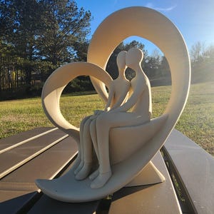 Puede incluir: Una escultura de cerámica blanca de dos figuras sentadas dentro de un marco en forma de corazón. La escultura está sobre una mesa de madera.