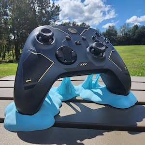 May include: A black and gold Xbox controller with a blue slime stand. The controller has a textured surface and gold accents. The stand is made of a soft, flexible material.