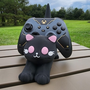 Pode incluir: Um suporte para controlador em forma de gato preto com orelhas cor-de-rosa e detalhes brancos. O suporte segura um controlador Xbox preto e dourado.