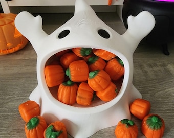 May include: A white ceramic ghost-shaped bowl filled with orange candy pumpkins. The bowl has a wide open mouth and two black eyes.
