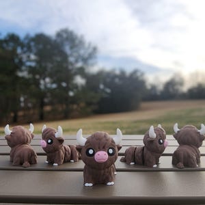 Puede incluir: Cinco figuritas miniatura marrones y blancas de un toro de dibujos animados con ojos grandes y una nariz rosa. Las figuritas están dispuestas en fila sobre una superficie marrón.