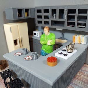 Puede incluir: Un juego de cocina en miniatura con una encimera gris, una estufa blanca, un refrigerador gris y gabinetes grises. Una figura en miniatura de una persona está de pie cerca del mostrador. Hay un pequeño pastel en el mostrador, y dos tazas y dos platos.