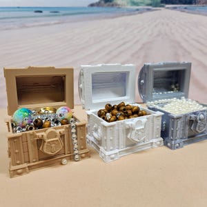 May include: Three open treasure chests filled with beads and jewels. The chests are tan, white, and gray, with decorative details. The tan chest contains iridescent and brown beads, the white chest has brown beads, and the gray chest is filled with white pearls.