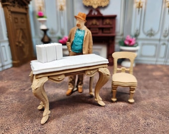 May include: A miniature dollhouse scene featuring a detailed desk with ornate legs, a stack of white books, and a small chair. A figurine of a man in a hat and coat stands nearby. The scene is set in a room with floral accents.