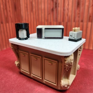 Op de afbeelding: Een miniatuurkeukenset met een wit aanrecht, drie kasten, een zwarte koffiezetapparaat, een zilveren magnetron en een zilveren broodrooster. Het aanrecht is versierd met een wit en grijs gespikkeld patroon.