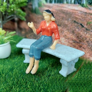 Könnte beinhalten: Eine Miniatur-Figur aus Plastik, die auf einer kleinen weißen Steinbank sitzt. Die Person trägt ein rotes Hemd und eine blaue Hose. Die Bank ist aus einem strukturierten weißen Material.