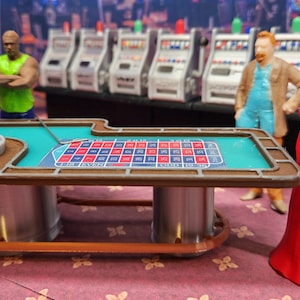 May include: A miniature roulette table with a green felt playing surface and a silver roulette wheel. The table has two silver legs and a brown wooden frame. There are three miniature figures standing around the table, including a woman in a red dress.