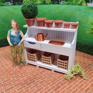 May include: A white wooden potting bench with a shelf and two drawers. The bench has five terracotta pots with plants, a copper watering can, and three brown wicker baskets.