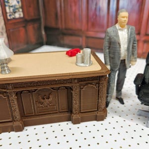 Puede incluir: Escritorio en miniatura marrón con tapa beige, una pequeña lámpara y un objeto rojo. Una silla de oficina negra y una figura de una persona con traje están cerca. El escritorio tiene tallas ornamentales.