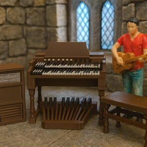 Peut inclure: Un orgue miniature marron avec un haut-parleur, un banc et une figurine jouant de la guitare. L'orgue a deux claviers et un pédalier. La figurine porte une chemise rouge et un jean bleu.