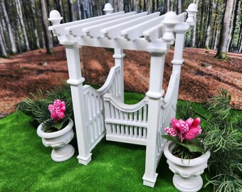 Puede incluir: Una pérgola blanca en miniatura con techo de listones y columnas decorativas. Dos urnas blancas con flores rosas flanquean la estructura, colocadas sobre un lecho de césped artificial verde. El fondo presenta una escena borrosa del bosque.