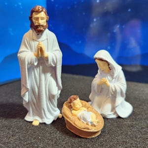 May include: A nativity scene set featuring three figurines: Joseph, Mary, and baby Jesus in a manger. Joseph stands with hands clasped, Mary kneels, and the baby lies in a brown cradle. All figures are dressed in white robes.