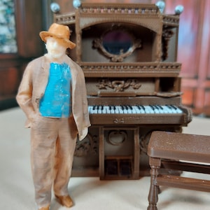 May include: A miniature figurine of a person wearing a brown hat and a blue vest standing next to a brown wooden miniature piano with a bench.