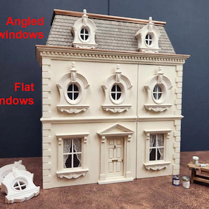 Op de afbeelding: Een crèmekleurig poppenhuis met een grijs pannendak, voorzien van twee schuine dakramen, drie ovale ramen op de tweede verdieping en twee rechthoekige ramen naast de voordeur. De tekst luidt Angled windows en Flat Windows.
