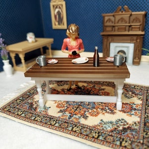 May include: A miniature dollhouse scene with a woman figurine standing at a wooden table with two plates, two mugs, and a bottle. The table is set on a cream-colored rug with a floral pattern.