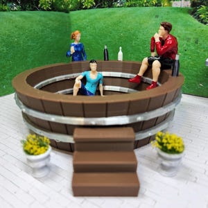Könnte beinhalten: Eine Miniatur-Whirlpool-Szene mit drei Figuren. Der braune Whirlpool hat silberne Bänder und Stufen, die hinaufführen. Kleine Topfpflanzen sind um den Whirlpool herum platziert. Der Hintergrund zeigt einen grünen Rasen.