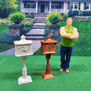 May include: Two miniature mailboxes, one white and one bronze, stand on a green surface. The white mailbox features a decorative relief. A miniature figure of a person stands nearby. The mailboxes are designed with a classic, ornate style.