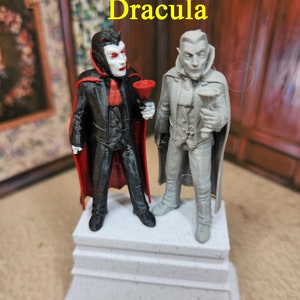 May include: Two 3D printed figurines of Dracula, one in black and red and the other in gray, standing on a white platform. The figurines are holding goblets.