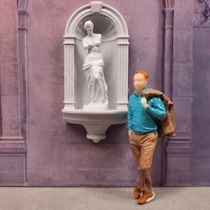 Puede incluir: Una figura en miniatura de una persona de pie frente a una fuente de pared blanca con una estatua de una mujer. La persona lleva una camisa azul y pantalones marrones. La fuente está decorada con un arco blanco y una base blanca.
