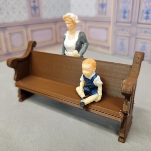 Könnte beinhalten: Eine Miniatur-Holzbank mit einer kleinen Figur eines Kindes in blauen Latzhosen, das darauf sitzt. Eine Frauenfigur in einer weißen Schürze und einem dunklen Kleid steht hinter der Bank. Die Szene spielt vor dem Hintergrund eines Raumes mit blau-weißen Paneelen.