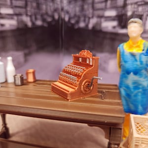 May include: A miniature wooden cash register with a copper finish. The register has a crank on the side and a display window with numbers. The register is sitting on a wooden table.