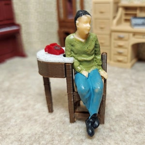 Può includere: Una figurina in miniatura di plastica di una persona seduta su una piccola sedia di legno con un telefono a disco rosso su un tavolo accanto a lei.