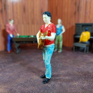 May include: A miniature figurine of a person playing a guitar. The figurine is wearing a red shirt and blue jeans. The figurine is standing on a brown surface.