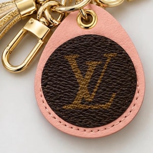 May include: A teardrop-shaped keychain featuring a light pink leather border surrounding a circular dark brown center with a gold LV monogram print. The accessory includes a gold-toned metal lobster clasp and a matching gold-toned tassel.