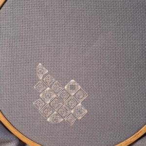 May include: A cross-stitch embroidery project with a geometric design. White diamond and square shapes are stitched onto blue-grey fabric. The fabric is held in place by a wooden embroidery hoop, highlighting the intricate needlework and the texture of the fabric.