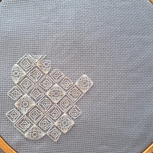 May include: A close-up of cross-stitch embroidery on grey fabric. The design features a geometric pattern of white diamonds and squares. The embroidery is partially framed by a wooden hoop, showcasing the craft's texture and detail.