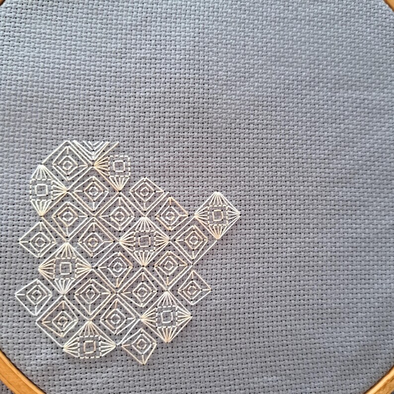 May include: A close-up of cross-stitch embroidery on grey fabric. The design features a geometric pattern of white diamonds and squares. The embroidery is partially framed by a wooden hoop, showcasing the craft's texture and detail.